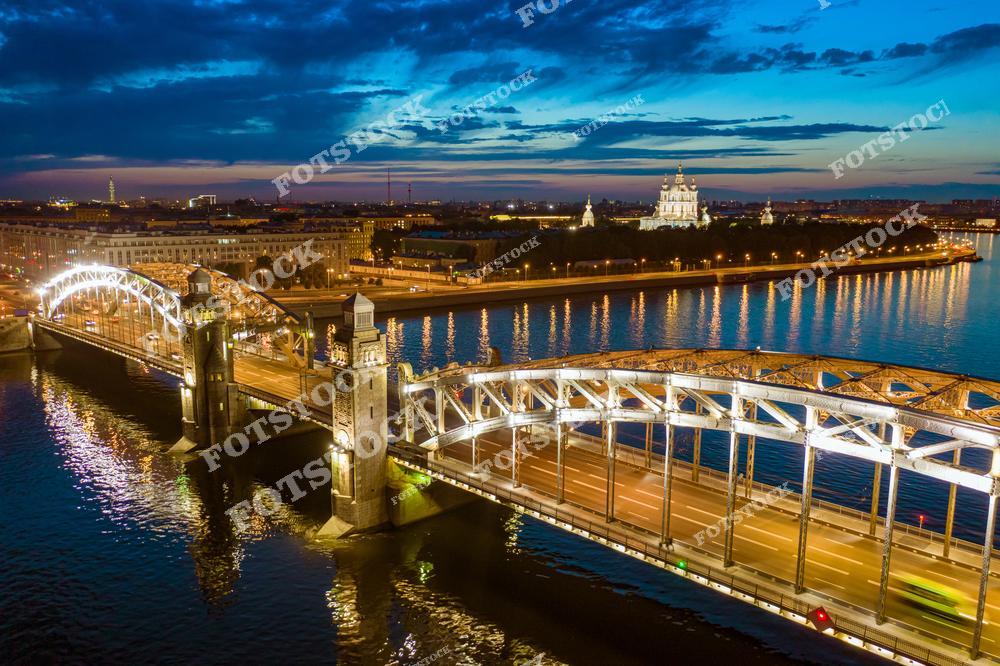 Фотография ночного Санкт-Петербург, Россия. На изображении показан Большеохтинский мост, также известный как мост Петра Великого, в эффектной ночной подсветке. Фотоснимок передаёт атмосферу вечернего города: огни иллюминации отражаются в воде, подчёркивая металлические конструкции моста и линии каналов Петербурга. Фото связано с темой речных прогулок и круизов по Санкт-Петербургу, популярных маршрутов для путешествий по Российской Федерации. Это изображение отражает романтику ночных видов и архитектуру мостов северной столицы.